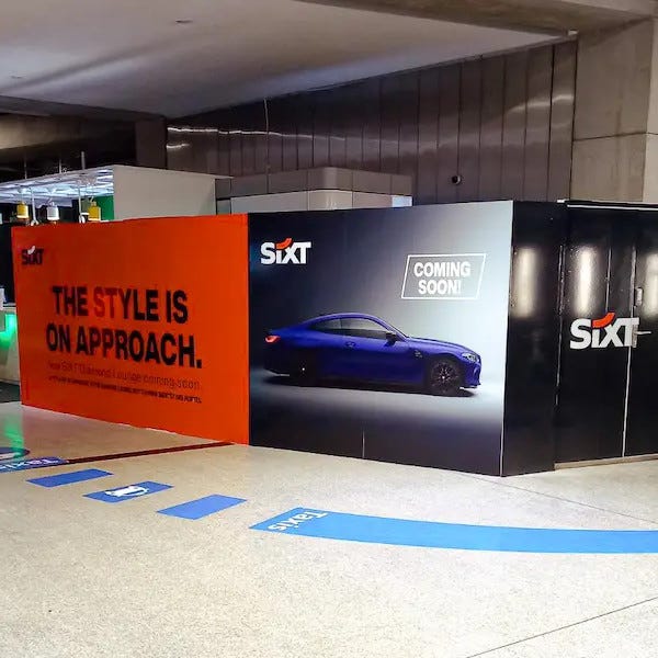 palissade de chantier aux couleurs de l'entreprise Sixt soit orange et noir avec une phrase écrite en noir a gauche et une voiture moderne en photo a droite