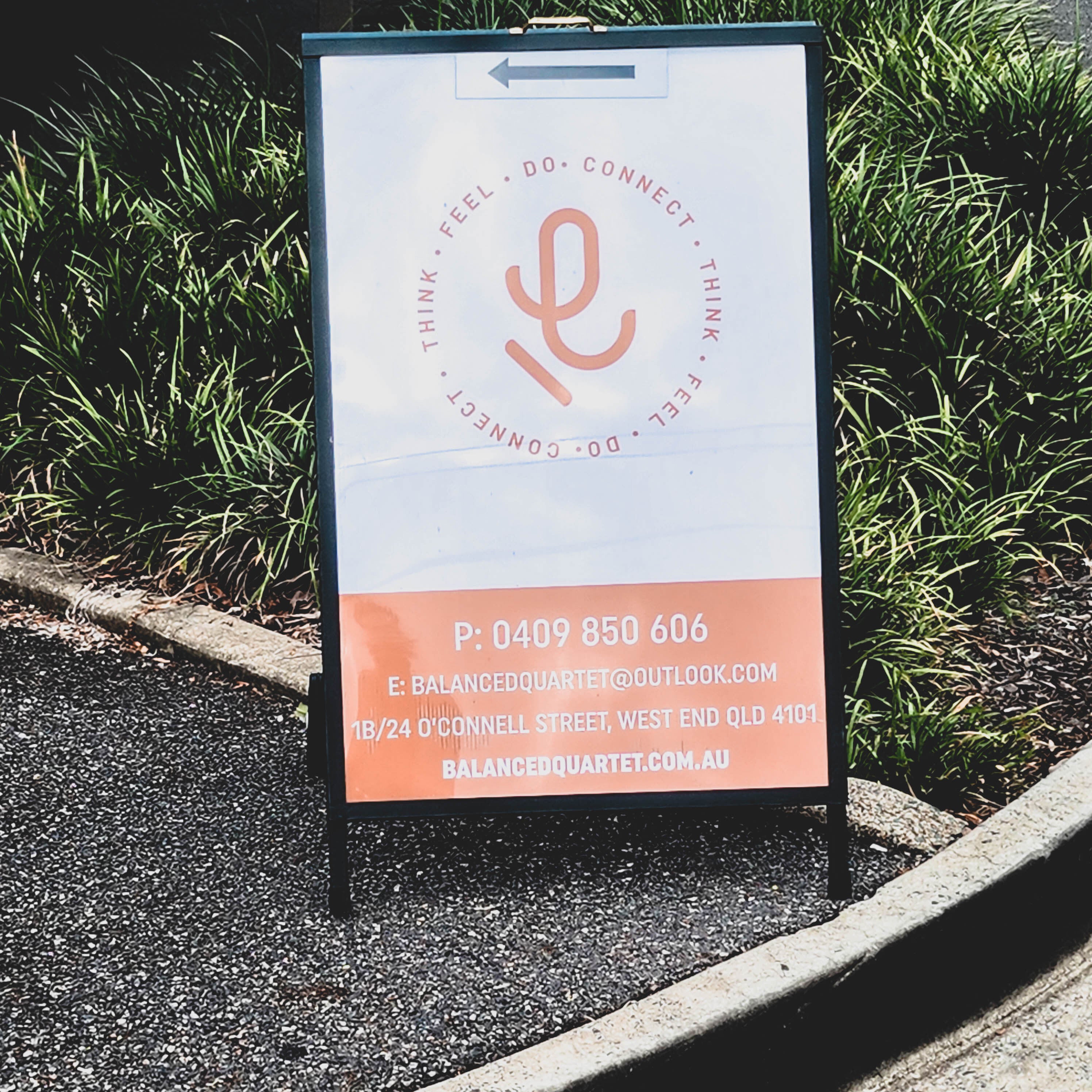A frame sign standing on pavement in front of green bush with white and red design comprising business logo and business details
