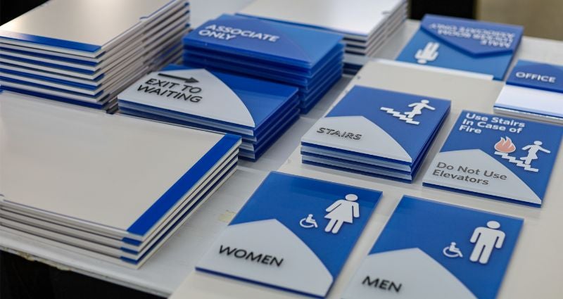 A collection of blue and white ADA-compliant signage featuring tactile lettering and universal pictograms for restrooms, stairs, and emergency exits.