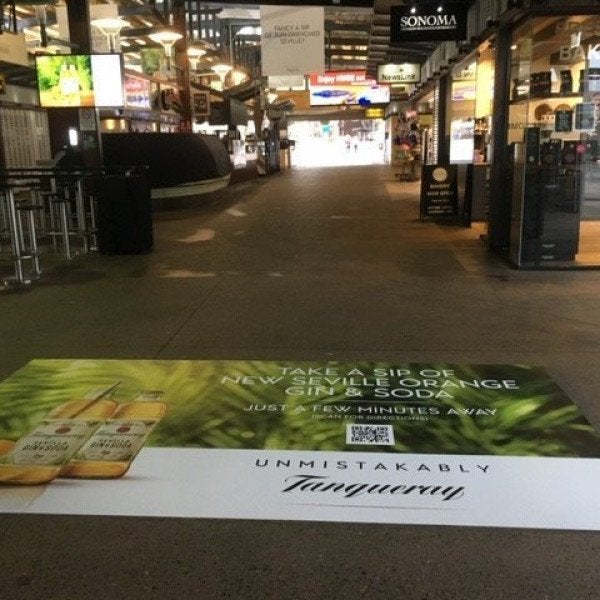 A floor graphic in a shopping centre that has a drink product advertisement with rainforest background and white branding
