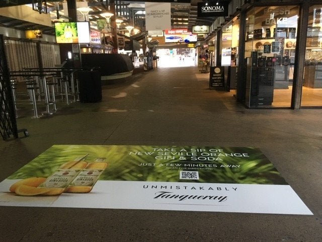 A floor graphic in a shopping centre that has a drink product advertisement with rainforest background and white branding