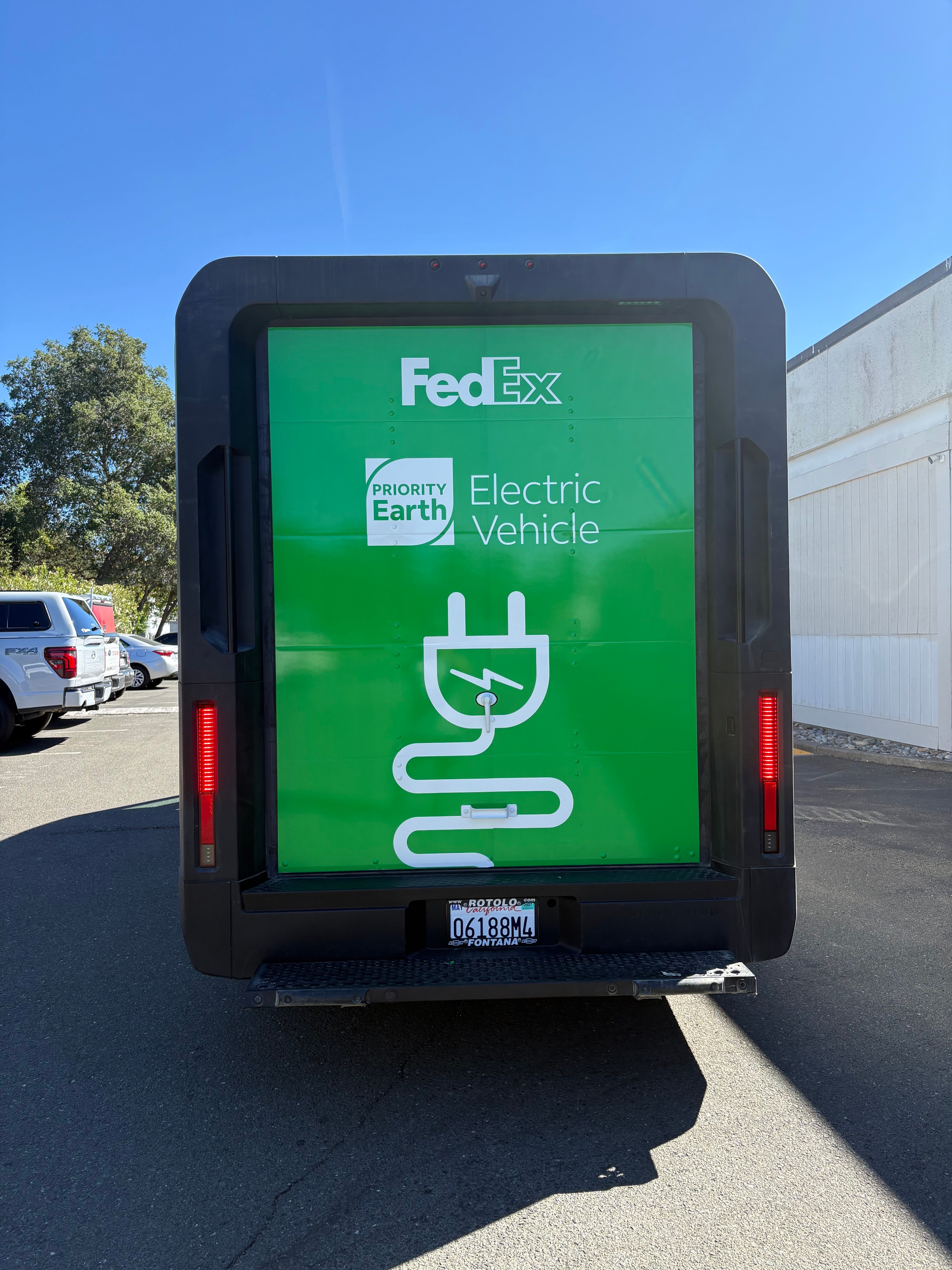 Rear view of a FedEx BrightDrop electric delivery van with green Priority Earth graphics and a plug icon.
