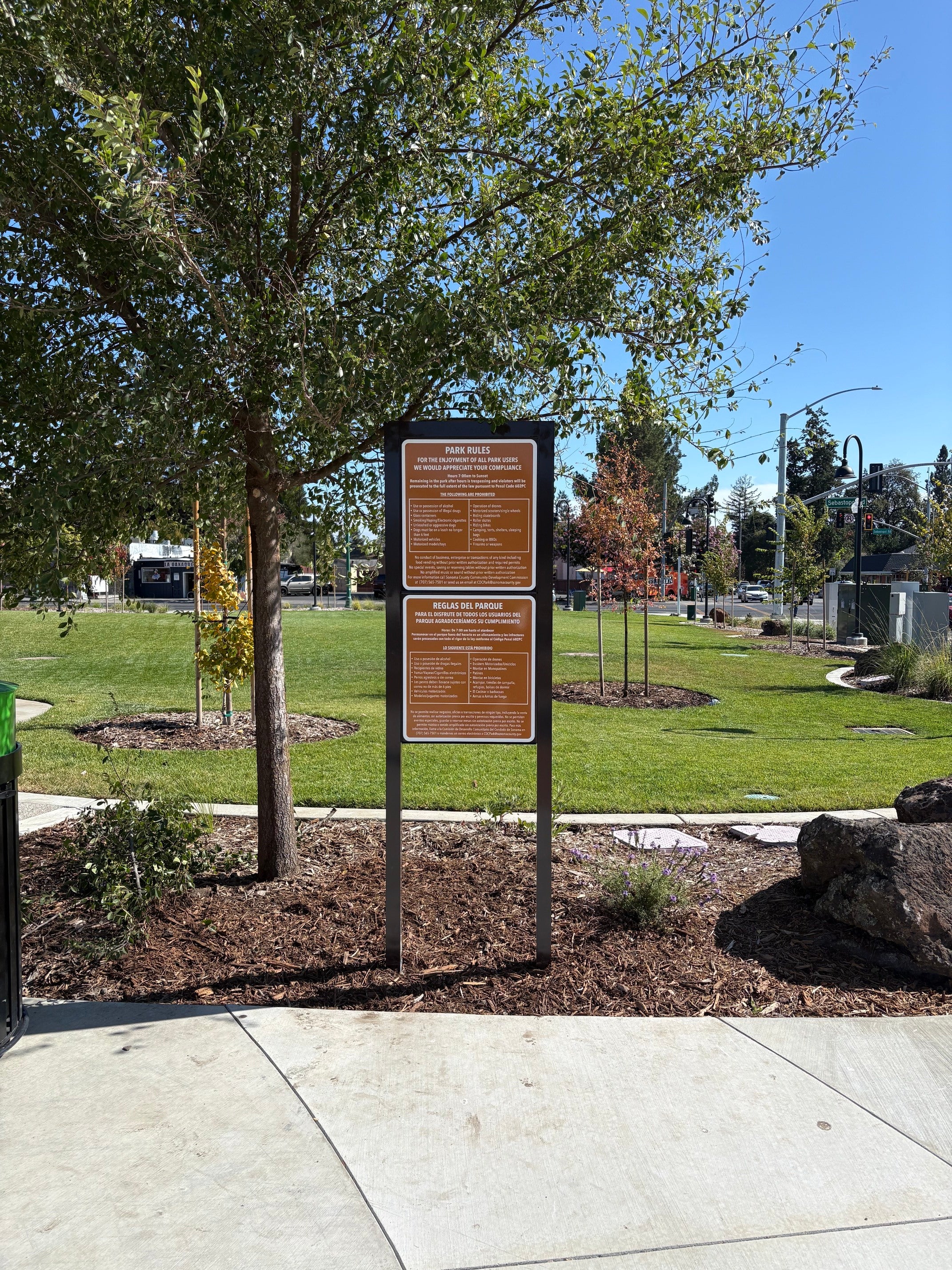 Freestanding bilingual park rules sign in a landscaped area at Plaza Permanente in Santa Rosa