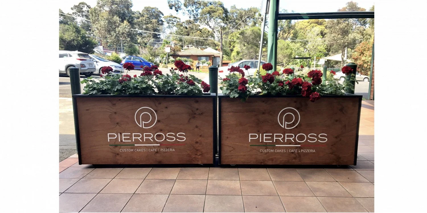 Thick timber cafe barriers with flowers planted in the top and with white branded elements on the timber