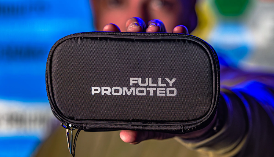 Close-up of a black branded pouch reading ‘Fully Promoted,’ held out toward the camera against a blurred background