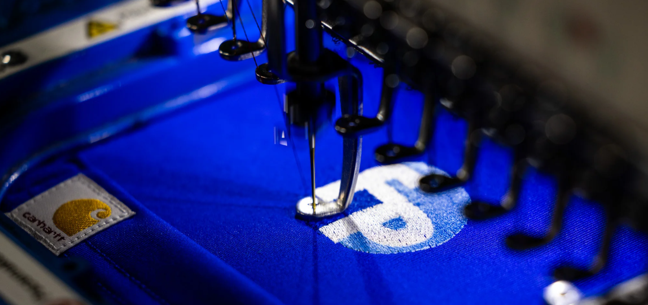 An industrial embroidery machine needle stitching the Fully Promoted circle logo onto bright blue fabric.